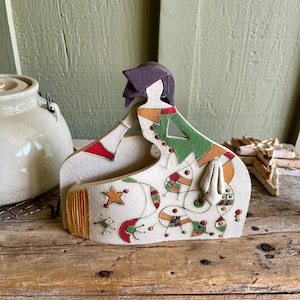 May include: A ceramic figurine of a stylized woman in a green, red, and yellow dress with geometric patterns. The figurine is painted with white, black, and brown accents.