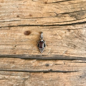 May include: A small, silver pendant with a rectangular, deep red gemstone at its center. The pendant has a detailed design with small, round accents and a loop for a chain. The pendant is displayed on a rustic, wooden surface.