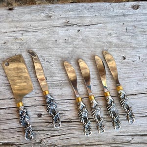 May include: A set of cheese knives and a cleaver with silver and gold handles. The handles are decorated with a grape vine design. The blades are gold and silver. The cleaver is rectangular and the knives have rounded blades.