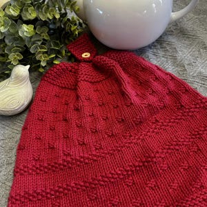 May include: A knitted red kitchen towel with a button fastening. The towel has a textured pattern and is designed to hang. A white teapot and decorative bird are in the background.