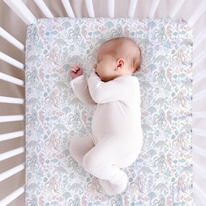 May include: A baby in a white sleepsuit lies on a cot sheet with a sea-themed pattern. The sheet features illustrations of octopuses, seahorses, and other marine life in soft pastel colours. The cot has white wooden slats.