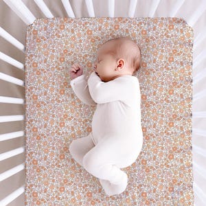 May include: A baby in a white onesie sleeps on a floral crib sheet. The sheet has a pattern of small orange and beige flowers on white. The cot has white wooden slats. The scene is viewed from above.