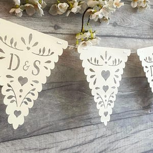 May include: A white paper bunting banner featuring triangular flags with intricate laser-cut patterns of hearts and foliage. One flag displays the letters D & S in a serif font. The flags are connected by a thin white string and hang horizontally.