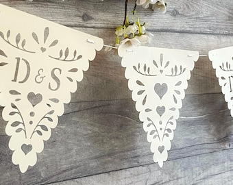 May include: A white paper bunting banner featuring triangular flags with intricate laser-cut patterns of hearts and foliage. One flag displays the letters D & S in a serif font. The flags are connected by a thin white string and hang horizontally.