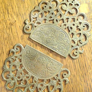 May include: Two matching semi-circular brass decorative plates featuring intricate openwork scroll patterns and embossed floral motifs. Each piece has a flat, textured central section with carved flowers and leaves surrounded by ornate filigree.