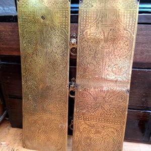 May include: Two antique brass door plates with intricate floral and geometric engravings. The plates have a rectangular shape with a decorative top, featuring sunburst and floral designs. The brass has a warm, aged patina, suggesting age and use.