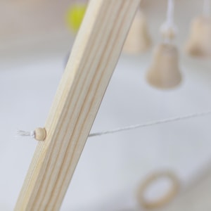 Op de afbeelding: Close-up van een houten babymobielframe met een wit touwtje bevestigd aan een houten kraal. Het touwtje is met een knoop aan het frame bevestigd.