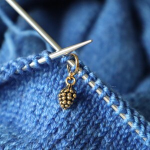 Op de afbeelding: Close-up van blauw gebreide stof met zilveren breinaalden en een goudkleurige steekmarkeerder. De steekmarkeerder heeft de vorm van een kleine dennenappel. De naalden zijn in de gebreide steken gestoken.