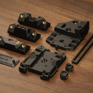 Puede incluir: Un conjunto de componentes negros impresos en 3D con inserciones de color lat&oacute;n, que incluyen formas rectangulares y geom&eacute;tricas. Una pieza tiene el texto "MARK EDGE REMOVE BEFORE CUTTING". Los elementos est&aacute;n dispuestos sobre una superficie de madera.