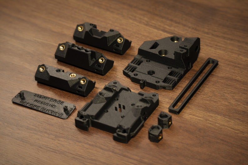 Puede incluir: Un conjunto de componentes negros impresos en 3D con inserciones de color lat&oacute;n, que incluyen formas rectangulares y geom&eacute;tricas. Una pieza tiene el texto "MARK EDGE REMOVE BEFORE CUTTING". Los elementos est&aacute;n dispuestos sobre una superficie de madera.