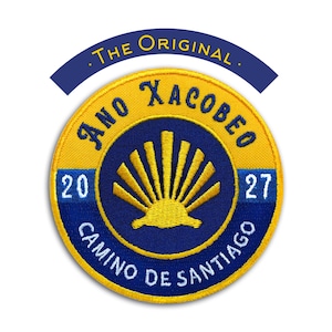 Puede incluir: Un parche circular con borde amarillo y centro azul. El parche presenta el texto "ANO XACOBEO CAMINO DE SANTIAGO" y los años "20" y "27". Un diseño de sol estilizado está en el centro, con las palabras "THE ORIGINAL" arriba.