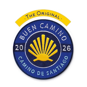 Puede incluir: Un parche azul redondo con las palabras "BUEN CAMINO" y "CAMINO DE SANTIAGO" en letras blancas. Un diseño de concha de vieira amarilla está en el centro. El parche también incluye los números "20" y "26". Encima del parche hay una pancarta amarilla con las palabras ".THE ORIGINAL."