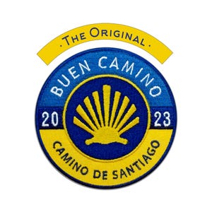 May include: A circular patch with a blue base and yellow detailing. The words "BUEN CAMINO" and "CAMINO DE SANTIAGO" are visible. The patch also features the text "2023" and ".THE ORIGINAL.". A yellow scallop shell design is in the centre.