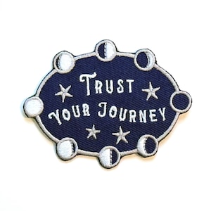 May include: A navy blue embroidered patch with the text "Trust Your Journey" in white script. The patch features a border of moon phases and stars, creating a celestial design. The patch is oval-shaped.