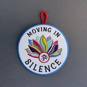 May include: Embroidered patch with a white background, blue border, and a red loop. The patch features a colorful lotus flower design and the text "MOVING IN SILENCE" in a circular arrangement. The Om symbol is in the center.
