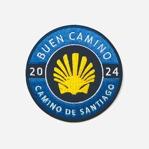 May include: Embroidered patch with a blue border and the words "BUEN CAMINO" and "CAMINO DE SANTIAGO" in white. The center features a yellow scallop shell and the year "2024".