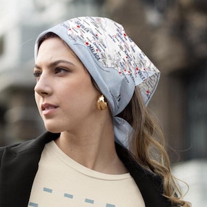 Puede incluir: Un pañuelo azul claro con un diseño de mapa, atado alrededor de la cabeza. El mapa incluye nombres de ciudades como Málaga y Valencia. El pañuelo se combina con pendientes de aro dorados y una camiseta color crema.