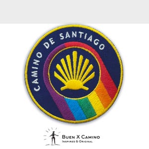 May include: A round, blue embroidered patch with a yellow scallop shell and a rainbow arc. The text "Camino de Santiago" is written in a circle around the shell.
