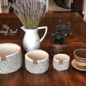 May include: Three crocheted baskets in various sizes with a colorful geometric pattern. The baskets are on a wooden table with a vintage coffee grinder, a pitcher of flowers, and a cup of coffee beans.