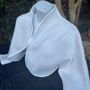 May include: A white, sheer bolero jacket with a collar and long sleeves. The jacket is displayed on a mannequin and has a delicate, textured appearance. The fabric appears to be lightweight and suitable for layering.