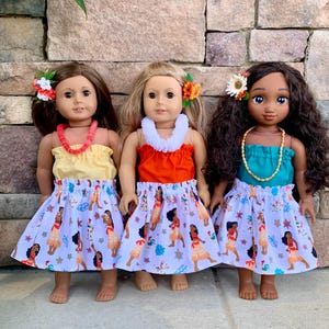 Pode incluir: Tr&ecirc;s bonecas vestidas com roupas com tema havaiano. Cada boneca usa um top colorido, uma saia com estampa de dan&ccedil;arina polin&eacute;sia e uma flor no cabelo. Elas tamb&eacute;m t&ecirc;m colares. As bonecas est&atilde;o em frente a uma parede de pedra.