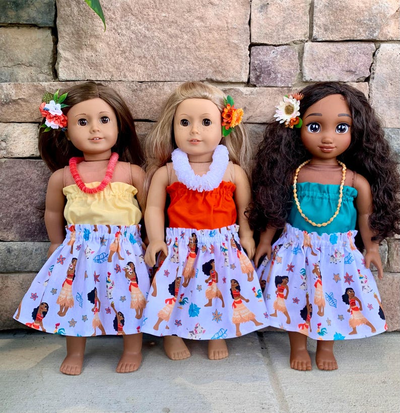 Pode incluir: Tr&ecirc;s bonecas vestidas com roupas com tema havaiano. Cada boneca usa um top colorido, uma saia com estampa de dan&ccedil;arina polin&eacute;sia e uma flor no cabelo. Elas tamb&eacute;m t&ecirc;m colares. As bonecas est&atilde;o em frente a uma parede de pedra.