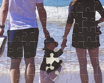 Peut inclure: Une famille de trois personnes debout sur une plage de sable, regardant l'océan. Le père porte un t-shirt blanc et un short noir. La mère porte un t-shirt noir et un short noir. L'enfant porte un t-shirt noir et une jupe rose et blanche.
