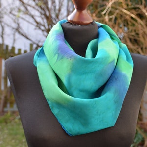 May include: A teal, green, and blue silk scarf with a tie-dye pattern is displayed on a black mannequin.