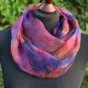 May include: A purple, pink, and orange silk scarf with a watercolor-like design. The scarf is draped around a mannequin's neck.