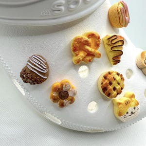 May include: A white shoe adorned with various food-themed charms. The charms include a pretzel, a pie, a cat face, a flower, a heart-shaped pastry, and a hot dog. The charms are in shades of brown, yellow, and pink.