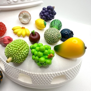 Op de afbeelding: Een witte schoen met een verscheidenheid aan kleurrijke fruitcharms eraan bevestigd. De charms omvatten een watermeloen, een ananas, een mango, een kokosnoot, een citroen, een kers, een tros druiven, een durian, een tros bramen en een bosbes.