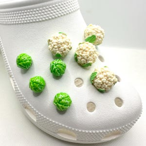 Puede incluir: Zapato blanco con varios dijes decorativos. Los dijes incluyen coles de Bruselas verdes y floretes de coliflor. Los dijes están adheridos al zapato, agregando un toque único y lúdico al calzado.