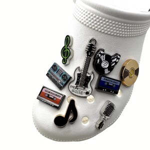 May include: White shoe adorned with music-themed charms. Charms include a guitar, cassette tapes, a treble clef, a microphone, and a vinyl record. The guitar charm has the text "NEW NOISE".