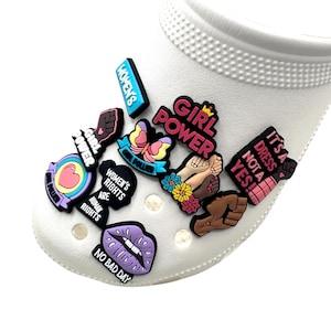 Può includere: Una scarpa bianca adornata con vari ciondoli colorati. I ciondoli presentano frasi come "Girl Power", "Women's Rights" e "It's a Dress Not a Yes!" insieme ad altri disegni grafici. La scarpa ha una superficie strutturata.