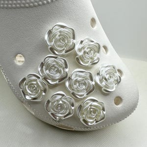 May include: A white shoe adorned with silver rose charms. The charms are clustered on the shoe's toe, adding a decorative touch. The shoe has multiple ventilation holes.