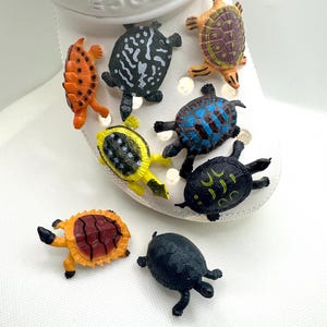 Puede incluir: Un conjunto de nueve figuritas de tortugas de plástico de colores. Las tortugas son de varios tonos de naranja, amarillo, verde, azul y negro. Todas son de diferentes tamaños y tienen diferentes patrones en sus caparazones.