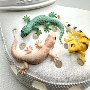 May include: A white Crocs shoe with four decorative lizard charms. The lizards are in shades of green, pink, yellow, and black and white. The green lizard has brown spots. The shoe has the Crocs logo on the side.