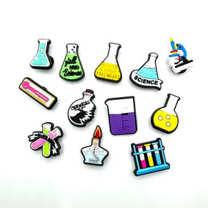 Puede incluir: Un conjunto de 12 amuletos de zapatos de colores con temática científica. Los amuletos incluyen vasos de precipitados, tubos de ensayo, un microscopio, una regla y un matraz con el texto "Science is magic that works".