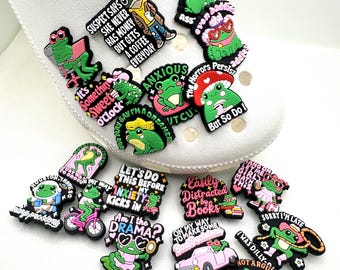 Pode incluir: Uma coleção de encantos de sapatos com tema de sapo colorido. Cada encanto apresenta um sapo verde com vários designs e frases como "Anxious" e "I'm Just Right". Os encantos são presos a um sapato branco.