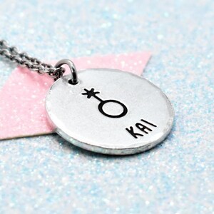 May include: A silver-toned circular pendant necklace featuring a stamped gender symbol and the name KAI in black lettering. The pendant is attached to a thin metal chain, showcasing a hammered texture on the surface of the round metal disc.