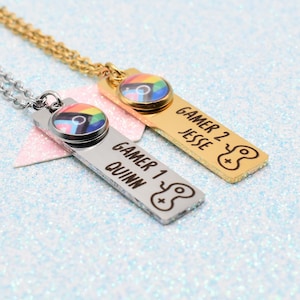 May include: Two gaming-themed pendant necklaces. One is silver, the other gold. Each has a rectangular pendant with a rainbow-coloured circular design and the words "Gamer 1 Quinn" and "Gamer 2 Jesse" engraved on them.