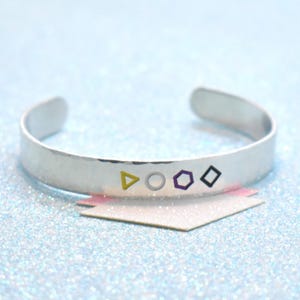 Op de afbeelding: Een zilveren manchetarmband met een gehamerde textuur. De armband heeft vier kleine, kleurrijke geometrische vormen: een gele driehoek, een witte cirkel, een paarse zeshoek en een zwarte ruit. De armband wordt gepresenteerd op een glinsterend blauw oppervlak.