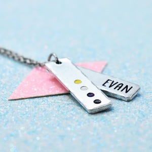 May include: A silver-toned metal pendant necklace featuring two rectangular tags. One tag is stamped with the name EVAN in black capital letters. The other tag displays four small, circular coloured accents in yellow, white, purple, and black.