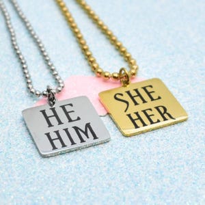 May include: Two square pendant necklaces with ball chains. One silver-toned pendant features the black text HE HIM, while the other gold-toned pendant features the black text SHE HER. Both pendants have a smooth, reflective metallic finish.