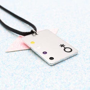 May include: Silver rectangular pendant necklace with a black cord. The pendant features a black symbol and four coloured dots: yellow, white, purple, and black. The necklace is on a light blue surface.