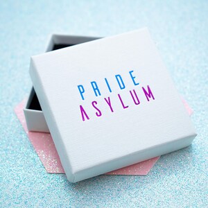 May include: A white box with the text "PRIDE ASYLUM" in blue and purple. The box is open, revealing a black interior. It rests on a pink, hexagonal shape against a sparkly, light blue background.