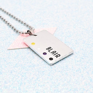 Op de afbeelding: Een zilverkleurige rechthoekige hangerketting met de naam "BLAIR" in zwarte letters. De hanger heeft vier gekleurde stippen: geel, wit, paars en zwart. De ketting is gemaakt van kleine, ronde, zilveren kralen.