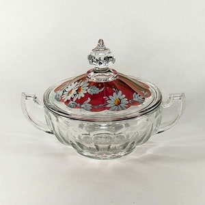May include: A clear glass sugar bowl with a lid featuring a red and white floral design. The bowl has a textured, faceted design and a knobbed lid.