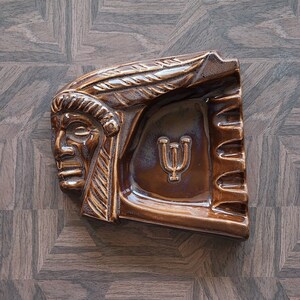 May include: A brown ceramic ashtray shaped like a Native American chief's head. The ashtray features a rectangular design with a recessed area for ashes and a symbol. The ceramic ashtray has a glossy finish and is brown in colour.