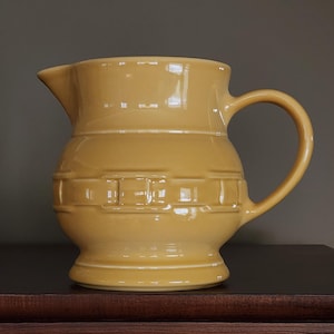 May include: A yellow ceramic pitcher with a woven design. The pitcher has a round body and a handle.